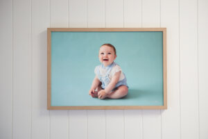 Oak framed canvas print of a baby sitting on a blue backdrop hung on a white vj groove panelling wall