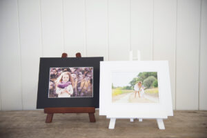 Two matted prints on a wooden bench, one has a black matt with an image of a girl resting on a wooden easel. The other is a white matted print of a girl and boy sitting on a white easel