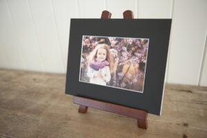 Black matted print of a girl with purple scarf witting on a wooden easel on a bench